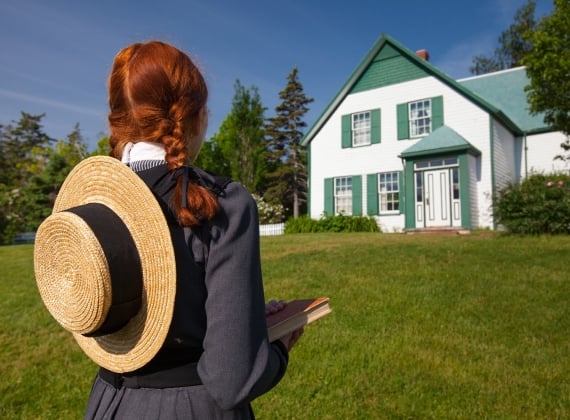 A girl with red hair in braids and a sun hat on her back looking at a white house on a sunny day.